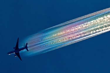 A commercial airplane in flight, leaving a bright, iridescent rainbow-colored contrail against a deep blue sky.