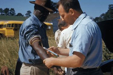 A man in a light blue shirt draws blood from the arm of a man wearing a hat, with a yellow school bus in the background.