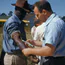 A man in a light blue shirt draws blood from the arm of a man wearing a hat, with a yellow school bus in the background.
