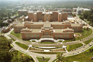 Aerial view of a large, multi-winged brick hospital complex surrounded by parking lots and wooded grounds.