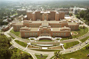 Aerial view of a large, multi-winged brick hospital complex surrounded by parking lots and wooded grounds.