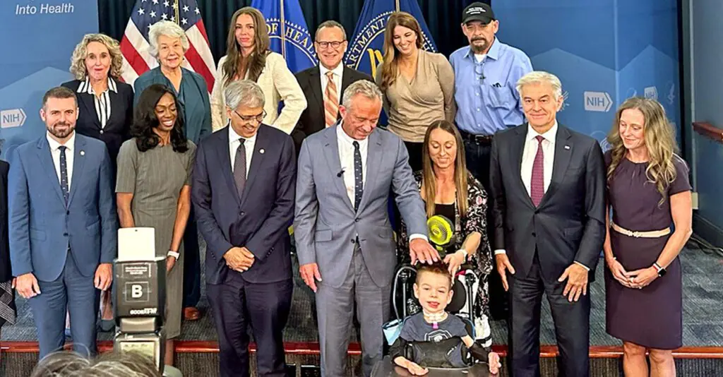 A group of adults stands behind a young boy in a wheelchair, with the man in the center placing a hand on the boy’s shoulder in front of National Institutes of Health (NIH) signage.